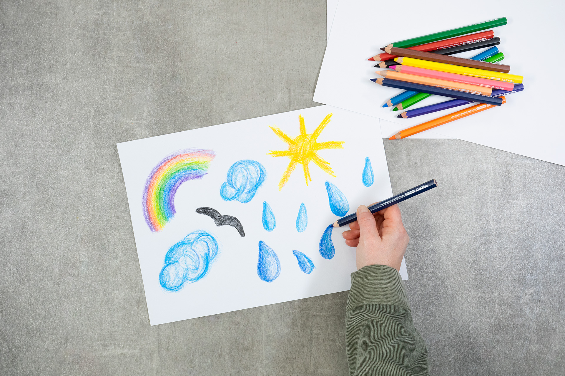 <p>Mit den leuchtenden Farben der KuKiMa Buntstifte male ich verschiedene Wetter-Symbole auf den weißen Bastelkarton. Regenbo